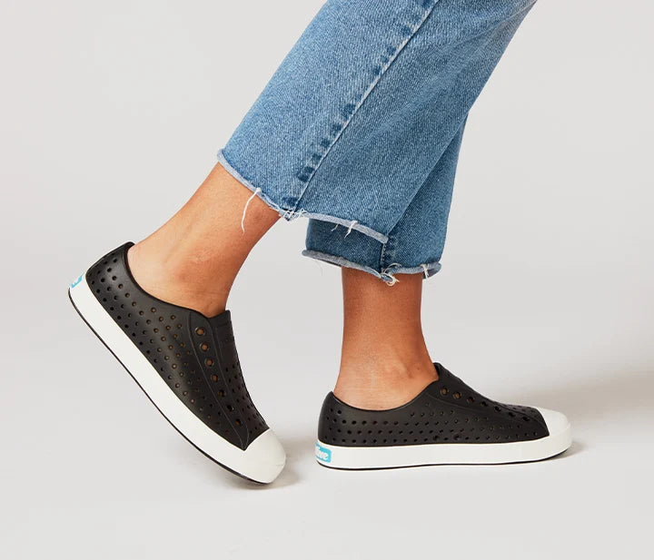 Black slip-on shoes with white soles worn with blue jeans on a light gray background
