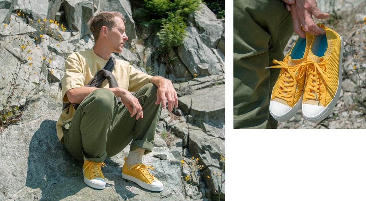 Guy wearing yellow trending sneakers crouches on rocky cliff. Guy holding yellow trending sneakers in hand.