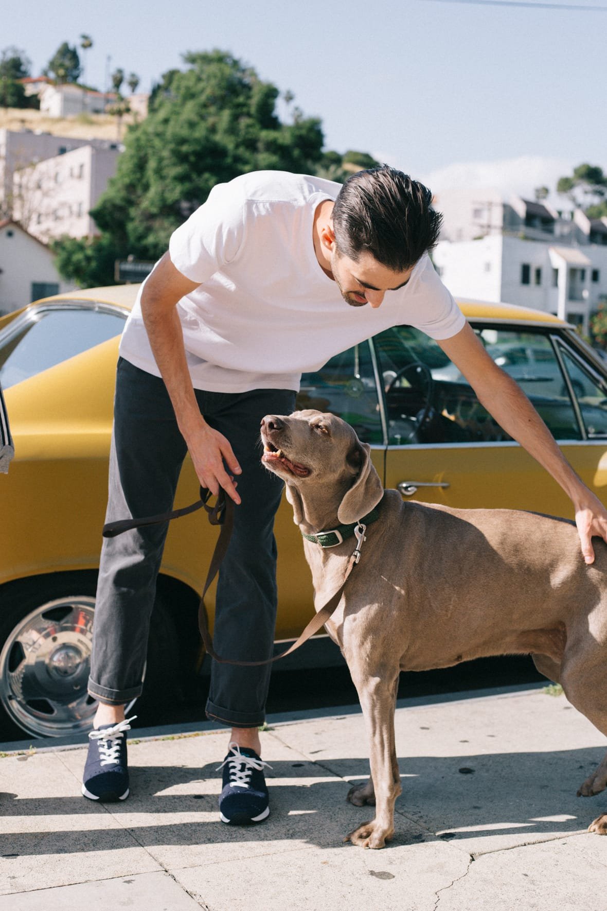 Guy wearing navy sneakers pets dog in front of yellow vintage car