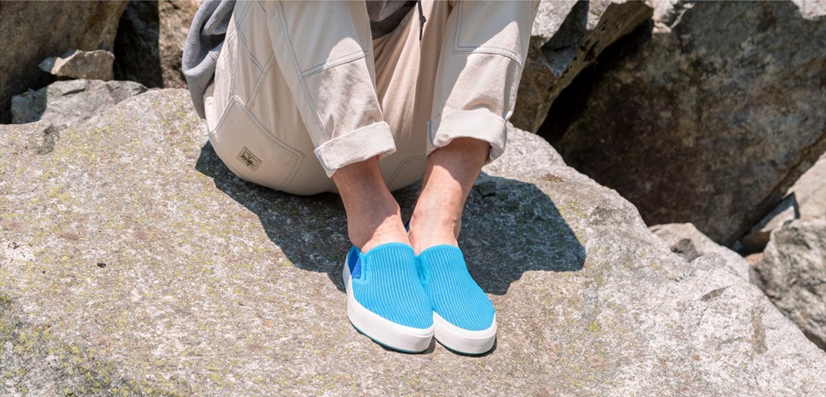 Person wearing blue slip-on stylish sneakers sitting on rock.