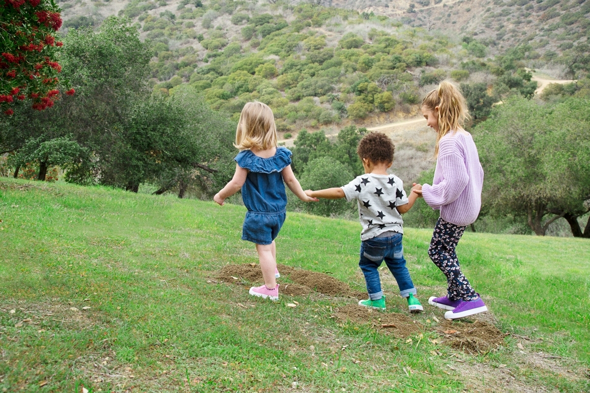 Three kids holding hands walk on a grass field.