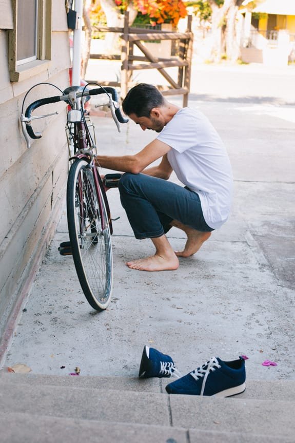 Guy crouching down to fix bike wheel in bare feet, with navy slip-on sneakers sitting nearby