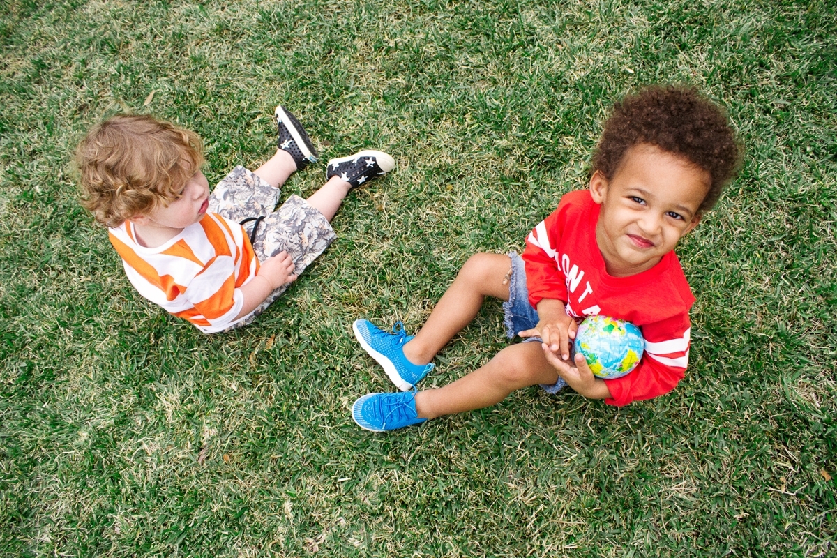 Two kids are sitting on a grass field wearing the Native Shoes Mercury Liteknit and the Jefferson in star print. One kid holds a globe.
