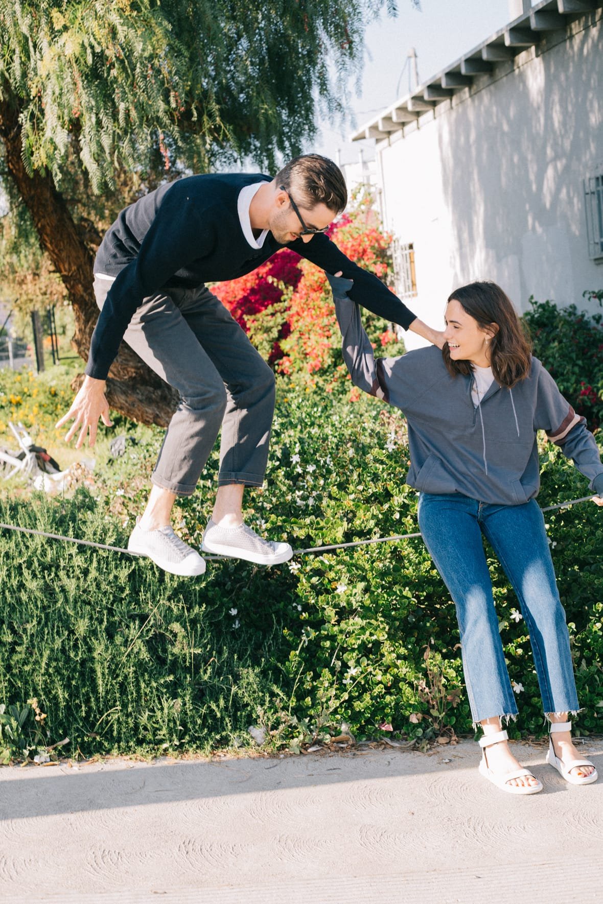 Guy balancing on suspended rope wearing grey sneakers holding shoulder of sitting girl who laughs while wearing white sandals