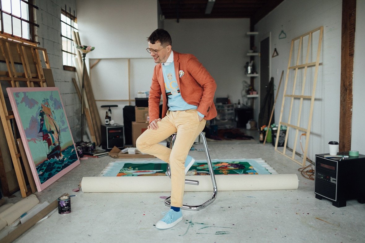 Andy Dixon laughing in his studio photographed on a stool wearing signature colors and blue Native Shoes.